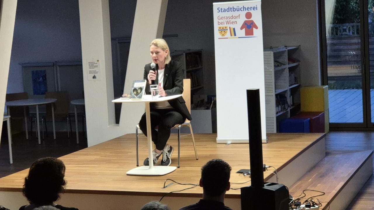 Claudia Rossbacher auf der Bühne von vorne, im Hintergrund das Logo der Stadtbücherei