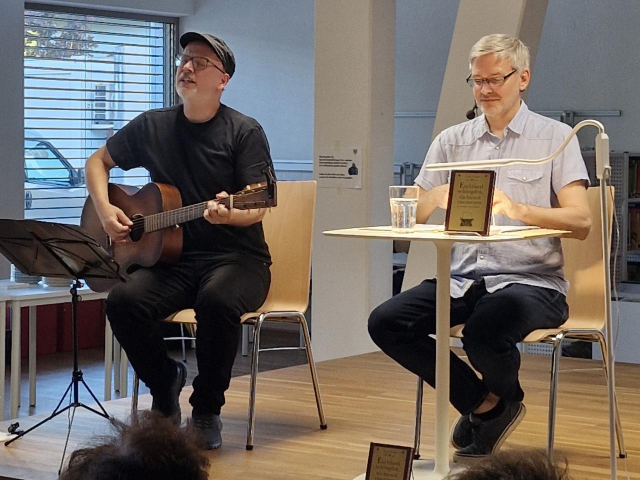 Helmuth Martin Wolf sitzt links auf der Bühne, spielt Gitarre und singt, daneben sitzt Autor Stefan Franke und hört zu