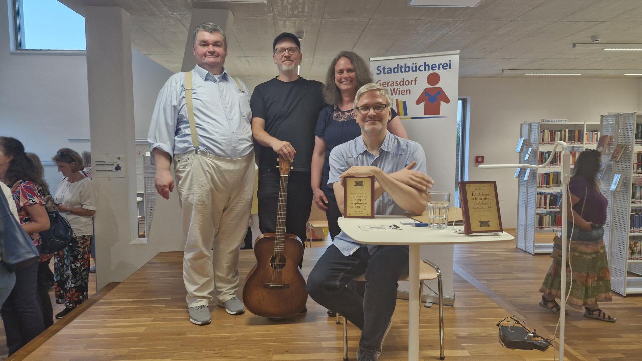 Gruppenbild - zu sehen 4 Personen, eine davon sitzt, die Gruppe befindet sich auf der Bühne im Hintergrund das Banner mit dem Logo der Stadtbücherei