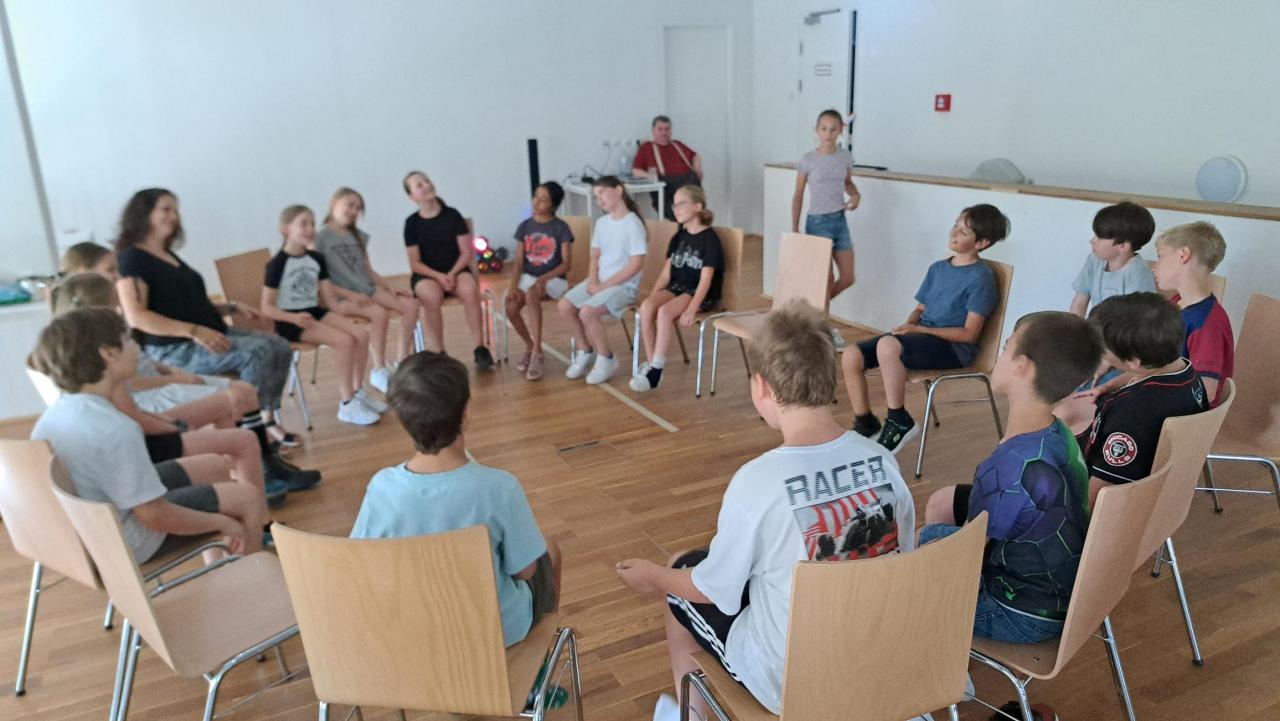 die Kinder und die Lehrerin sitzen auf Sesseln im Kreis und spielen das Spiel "Detektiv"
