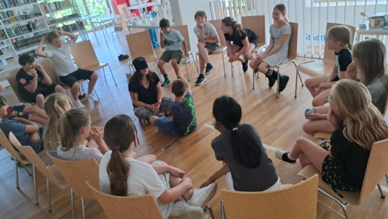 die Kinder sitzen im Kreis auf Sesseln, in der Mitte sitzt die Lehrerin mit verbundenen Augen und davor ein Kind, sie muss erraten wer vor ihr sitzt