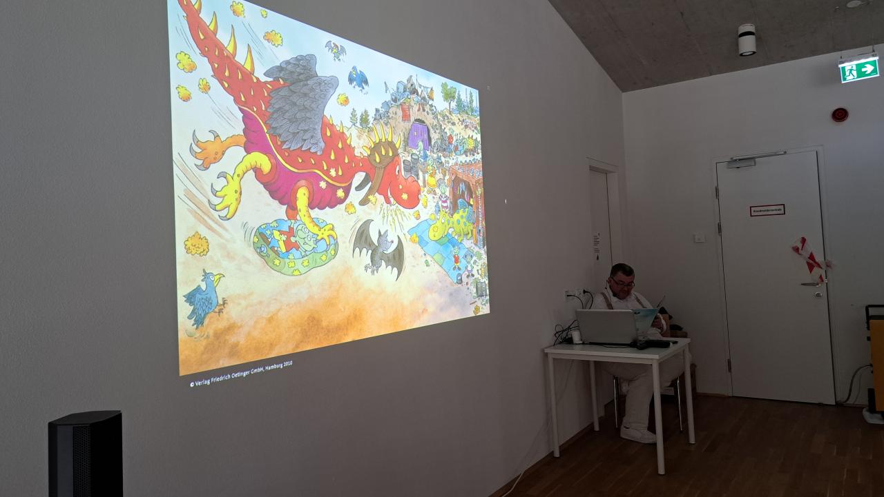Christian sitzt beim PC und liest die Geschichte vor, die Bilder werden mittels Beamer an die Wand projiziert - man sieht einen roten Drachen vor der Landung