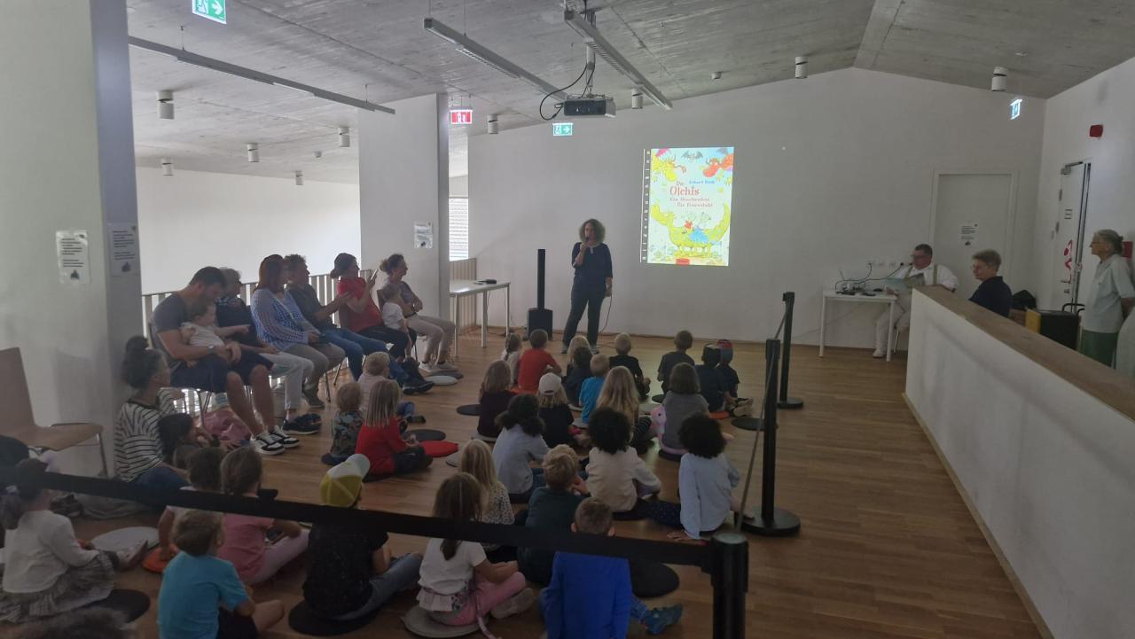 Kinder sitzen am Boden, Erwachsene am Rand auf Sesseln, vorne steht die Büchereileiterin und begrüßt 