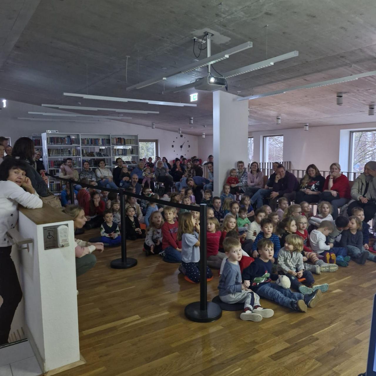 zahlreiche Kinder sitzen am Boden, Erwachsene runherum auf Sesseln