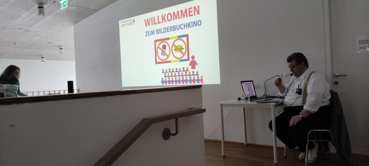 ein Mann sitzt am Tisch und spricht ins Mikrofon, an der Wand ist ein Bild, link steht eine Frau