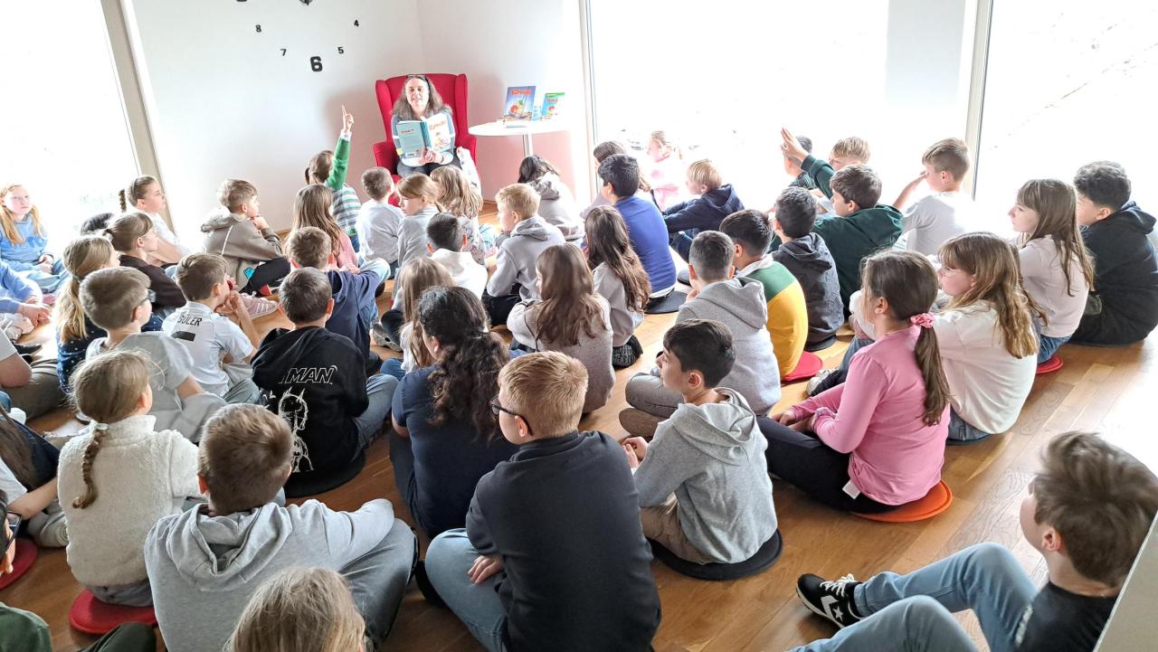 zahlreiche Kinder im Vordergrund, im Hintergrund eine Frau, die ein Buch liest