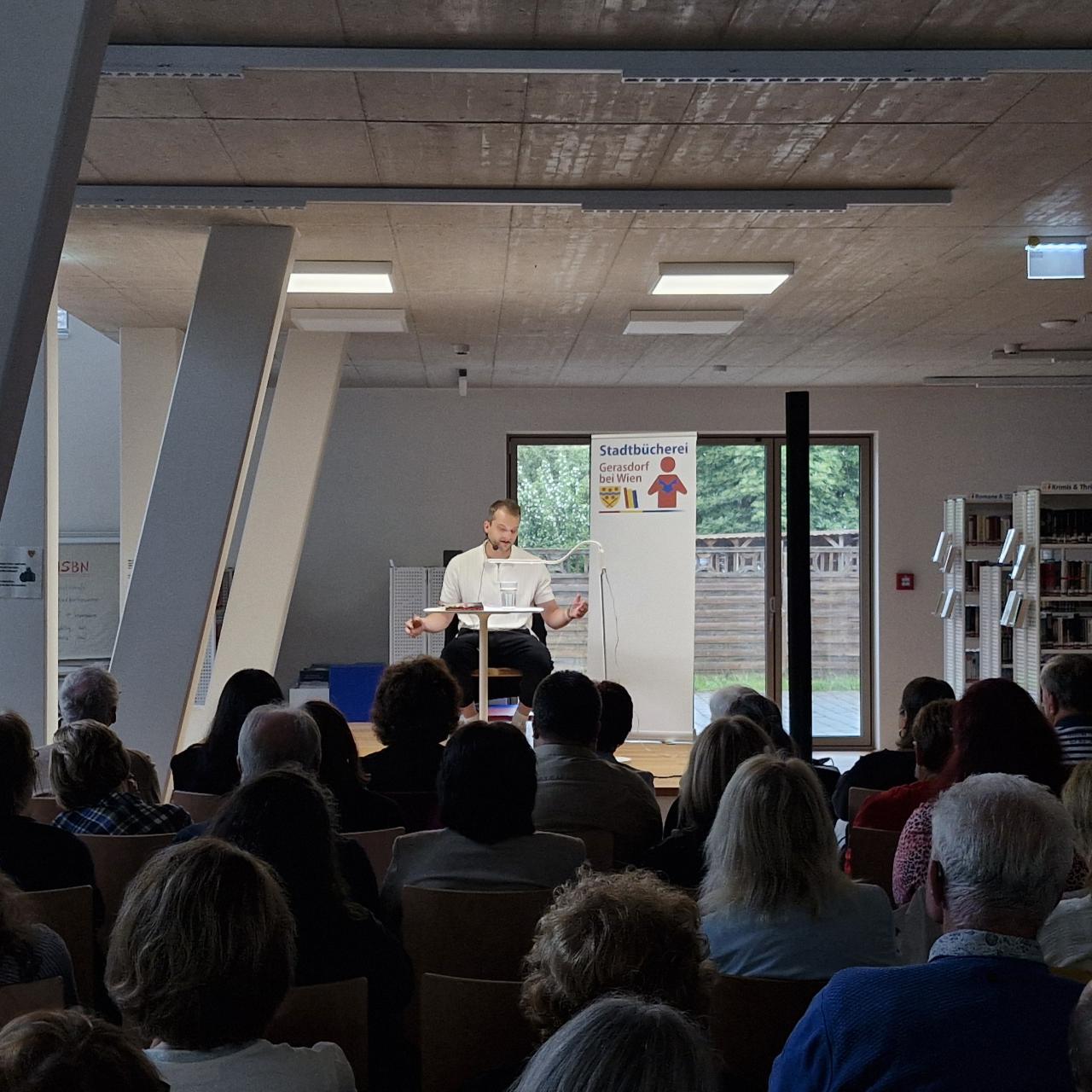 man sieht den Nachwuchsautor Lukas Reiff auf der Bühne sitzend und lesend, davor im Halbdunkeln das Publikum