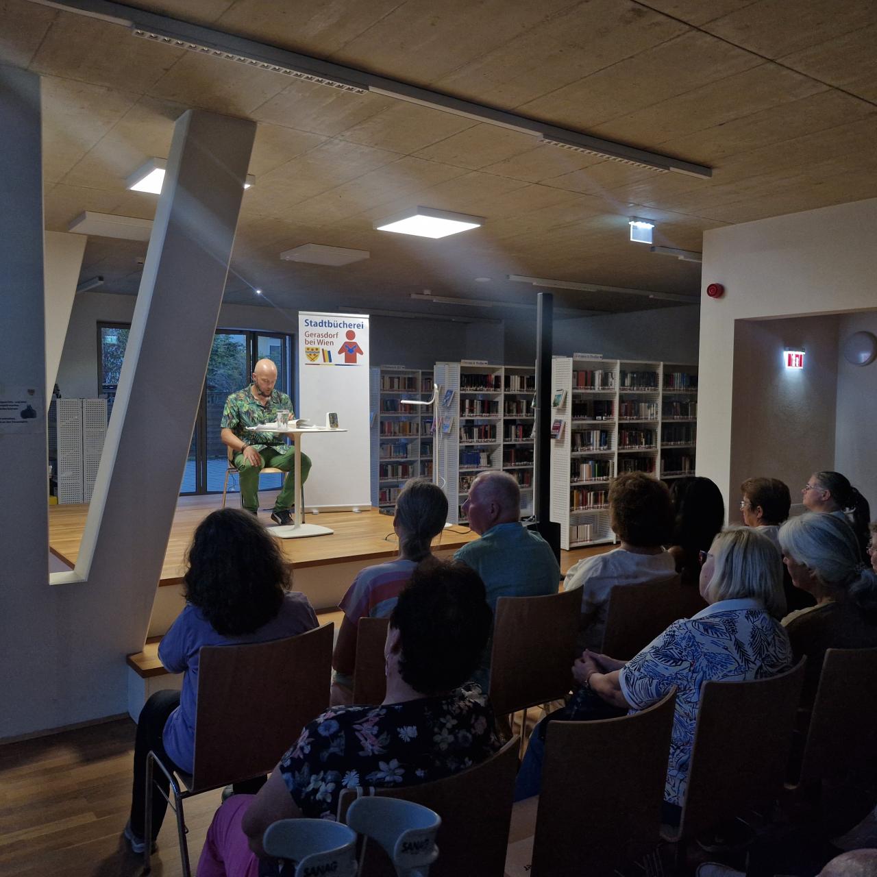nun sitzt der Autor auf der Bühne und liest aus einem Buch vor, vor der Bühne sitzen Menschen und hören zu