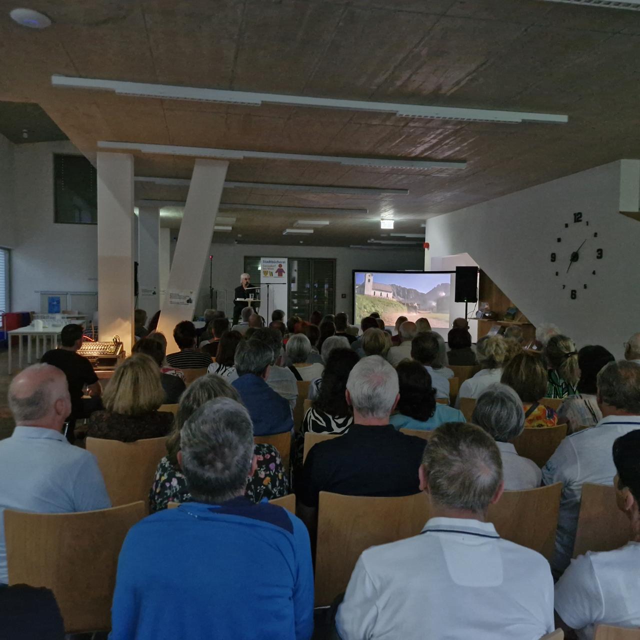 im Vordergrund zahlreiche Menschen auf Sesseln sitzend, im Hintergrund die Bühne mit dem Künstler, rechts vorne eine Leinwand, auf der ein Bild zu sehen ist